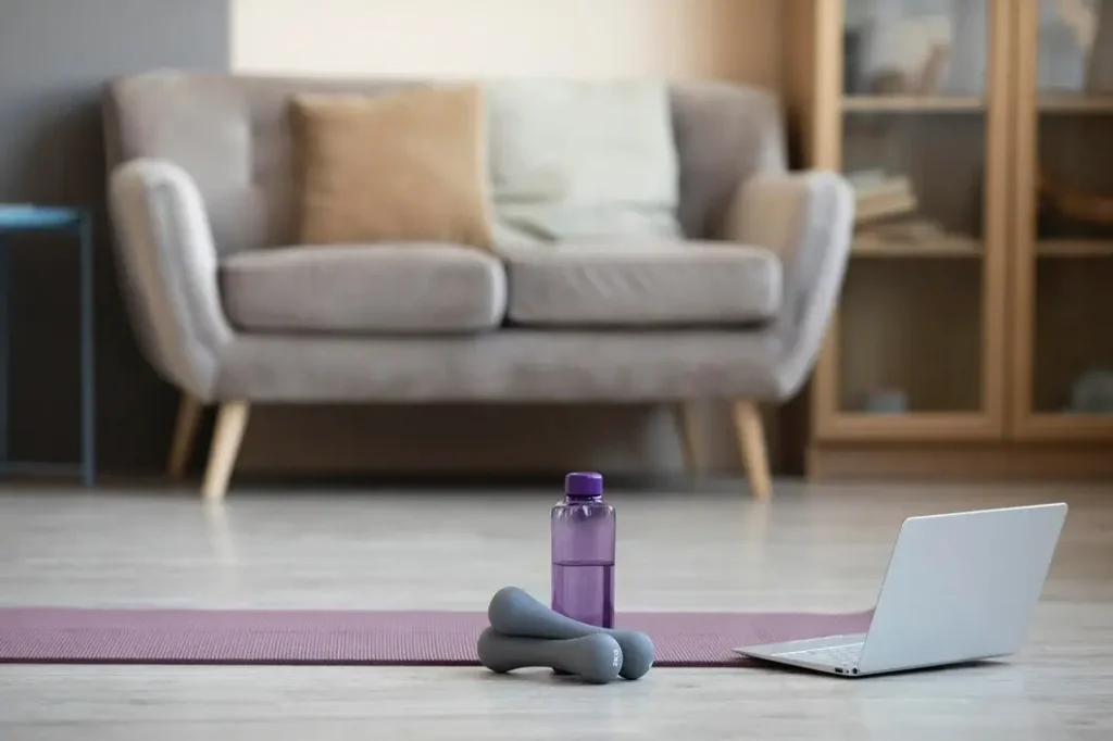 Yoga mat with dumbbells, water bottle, and laptop in a small space gym setup.