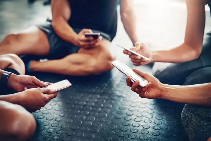 Group of gym-goers sitting in a circle using smart fitness tracker apps on their phones to share progress and stay connected.
